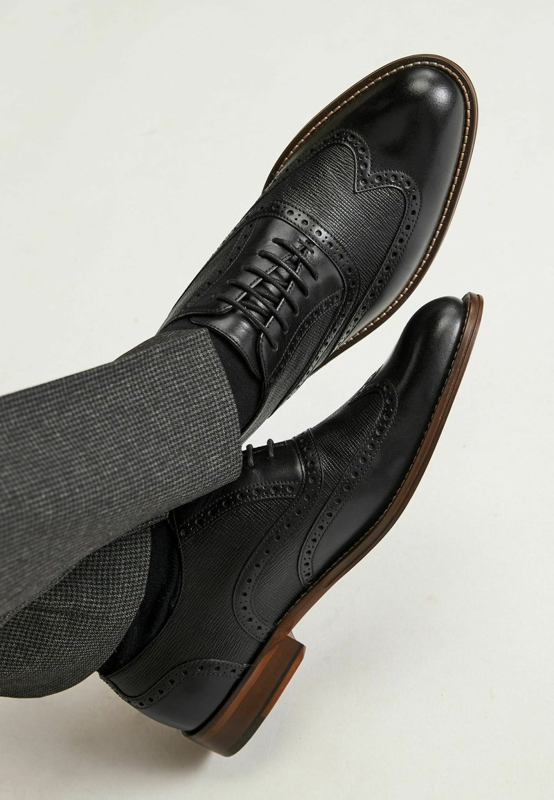 Chaussures de ville noires en cuir avec des détails en brogue, des côtés texturés et des bouts ronds. La semelle est d'un brun moyen avec une finition lisse.