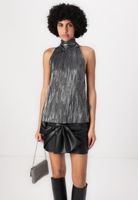 Metalni halter top s vertikalnim prugama, u kombinaciji s crnom suknjom od umjetne kože koja ima veliki detalj mašne. Model drži malu srebrnu torbicu.