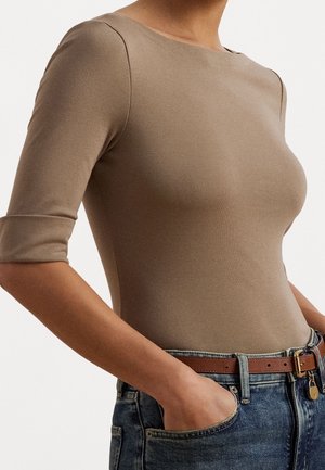 Vrouw die een aansluitend taupekleurig topje met driekwartmouwen draagt, gestopt in een blauwe spijkerbroek met een bruine leren riem en haar hand in de zak.