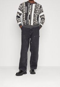 Gebreide trui in grijze en zwarte patronen, gecombineerd met losse zwarte spijkerbroeken met zakken en zilveren accenten, afgewerkt met zwarte schoenen.