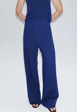 Persona con pantalones azules de corte holgado y una camiseta azul a juego, de pie con las manos detrás de la espalda sobre un fondo liso.