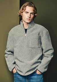 Sivý fleecový pulóver s priečnym zipsom, ktorý má predné vrecko a rebrované manžety. Nosí sa s modrými riflami na neutrálnom pozadí.