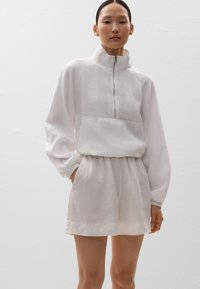 Weißer Leinenpullover mit langen Ärmeln, hohem Kragen und halbem Reißverschluss, kombiniert mit passenden Leinen-Shorts. Weiche Textur und lässige Passform.