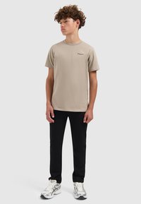 Beige katoenen T-shirt met korte mouwen en ronde hals, voorzien van een klein zwart logo op de borst, gecombineerd met zwarte broek en zilveren sneakers.