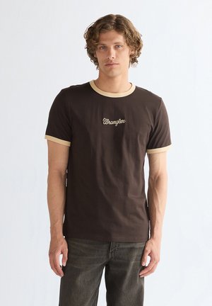 Młody mężczyzna o kręconych włosach, ubrany w brązowy t-shirt z beżowym obszyciem i tekstem "Wrangler", stoi na jednolitym białym tle.