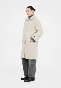 Donna in cappotto di lana beige di lunghezza media con dettagli sui bottoni, maglione grigio a collo alto, jeans neri lavati e stivaletti neri alla caviglia, in piedi di lato.