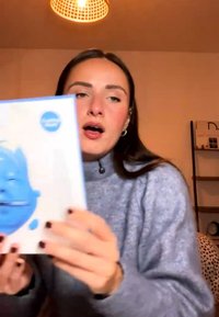 Kvinde i grå sweater holder og viser en blå og hvid bog med et illustreret ansigt på omslaget og taler i et varmt oplyst rum.