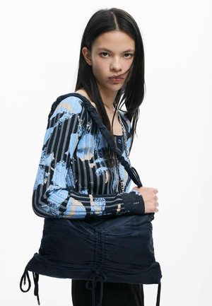 Must kangast kott, millel on kogutud disain ja nöörkinnis, hoitakse käes. Mudel kannab sinist mustrilist särke triipude ja laikudega.