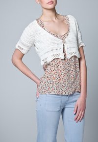 Bílý háčkovaný krátký kardigan se zvlněným lemem a zapínáním na knoflíky, vrstvený přes květovanou halenku a světle modré kalhoty.