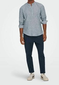 Jasnoniebieska lniana koszula z długim rękawem i okrągłym kołnierzykiem, zestawiona z ciemnoniebieskimi dopasowanymi spodniami i beżowymi sneakersami.