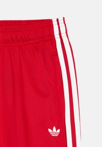 Κόκκινο αθλητικό παντελόνι με ελαστική μέση, με δύο λευκές ρίγες στα πλευρά και ένα μικρό λευκό λογότυπο στο κάτω αριστερό μέρος.