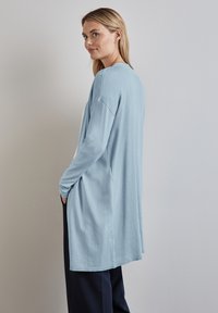 Ljusblå långärmad topp gjord av mjukt tyg, med avslappnad passform, neddragna axlar och sidofickor. Enkel halsringning och ribbstickade ärmslut.