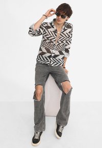Camicia a maniche lunghe a righe bianche e nere con un design geometrico, abbinata a jeans grigi strappati e sneakers nere. Gli accessori includono occhiali da sole.