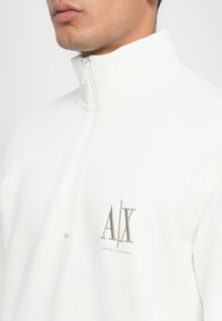 Vit halvdraget sweatshirt tillverkad av mjukt material, med en ton-i-ton "A/X"-logotyp broderad på vänster bröst. Slät textur, minimalistisk design.
