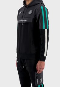 Sudadera negra con franjas turquesas y blancas en las mangas, bolsillo frontal y logo. El material es suave y tiene un diseño ajustado. Joggers negros con bolsillo con cremallera.