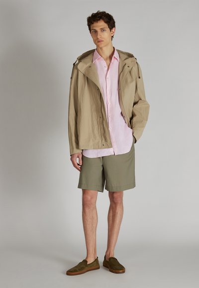 Joven de pie vistiendo una chaqueta con capucha beige, una camisa de botones rosa claro, pantalones cortos verde olivo y zapatos de slip-on marrones contra un fondo gris.