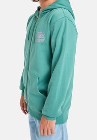 Personne portant un sweat à capuche zippé bleu sarcelle avec poche kangourou et logo Quiksilver rose sur la poitrine, assorti à un jean bleu clair.