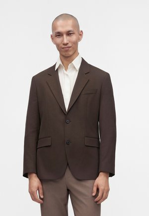 Mann trägt einen dunkelbraunen Blazer über einem weißen Hemd mit Kragen und hellbraune Hose, steht vor einem weißen, einfarbigen Hintergrund.