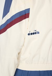 Krems krāsa jaka ar zilu un bordo svītrām, elastīgu jostu un izšūtu "diadora" logotipu kreisajā piedurknē. Gluda tekstūra.