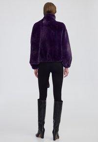 Donna che indossa un giubbotto in pelliccia sintetica viola, leggings neri e stivali in pelle nera al ginocchio, in piedi contro uno sfondo chiaro uniforme.