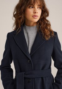 Kvinna med vågigt brunt hår, iförd en marinblå bälteullrock över en grå polotröja, som ser åt sidan mot en beige bakgrund.