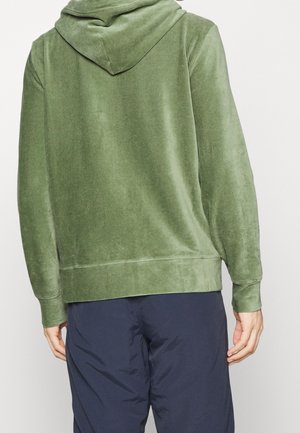 Persoon gekleed in een groene corduroy hoodie en marineblauwe broek, van achteren getoond tegen een eenvoudige witte achtergrond.