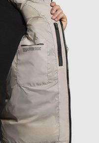 Svetlo siva pufasta jakna z gladko, sijočo zunanjostjo. Ima stranske žepe, črn zadrgo in natisnjeno besedilo na notranji podlogi.