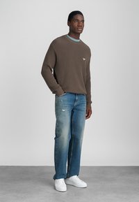 Rjav rebrast pulover z svetlo modrim ovratnikom, v kombinaciji z zbledelimi modrimi ravnimi hlačami in belimi supergami, stoji na nevtralni ozadju.