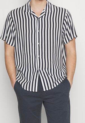 Homme portant une chemise à manches courtes noire et blanche à rayures verticales avec un pantalon sombre, les mains dans les poches.