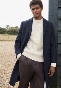 Abrigo de lana navy sobre un suéter de punto beige claro con un patrón texturizado, combinado con pantalones marrón oscuro. Presenta un cuello clásico y bolsillos.