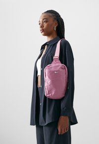Rosa Nike skulderveske med tekstiloverflate, glidelås-lukking og justerbar stropp. Båret med svart skjorte og hvit topp.