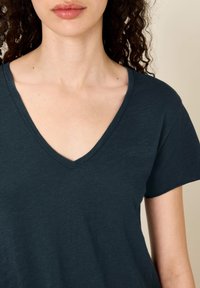 Žena s kudrnatými vlasy oblečená v tmavě modrém tričku s krátkým rukávem a výstřihem do V před jednoduchým světlým pozadím.