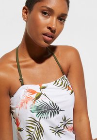 Costume da bagno bianco con motivi floreali e foglie tropicali verdi e arancioni, dotato di bretelle regolabili color verde oliva e una texture liscia e elastica.