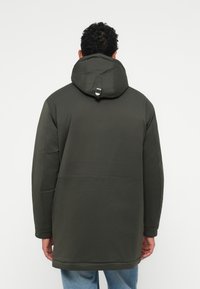 Sötétzöld parkajacket kapucnival és ezüst színű részlettel a hátán. Simaságot és textúrált anyagot kombináló, laza szabású.