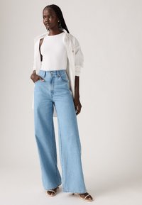 Woman with braided hair wearing a white sleeveless top, unbuttoned white shirt, high-waisted wide-leg blue jeans, and beige heeled sandals.