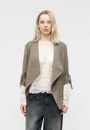 Blond kvinne med sølv hoops, en beige åpen jakke med draperinger over en hvit bluse i blonder, og mørkegrå høytlivsjeans.