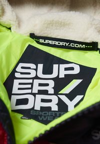Indersiden af Superdry-jakke med lys limegrøn foer, sort logo med fede hvide bogstaver og blød sherpa-lignende kant.