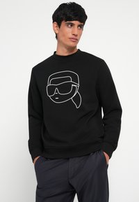 Felpa nera con una linea bianca che rappresenta un volto con grandi occhiali da sole. Il design presenta un classico collo a giro e polsini a costine.