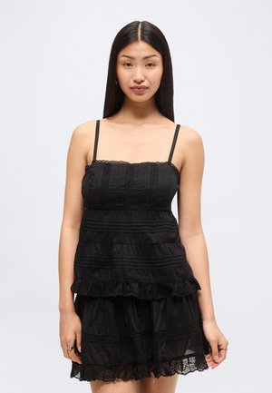 Νεαρή γυναίκα με μακριά μαύρα μαλλιά που φοράει ένα αμάνικο μαύρο φόρεμα με δαντέλα, στέκεται μπροστά σε λευκό φόντο.