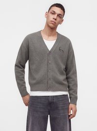 Ung mand iført en grå knap-op cardigan over en hvid skjorte og mørkegrå jeans, stående mod en ensfarvet lys baggrund.