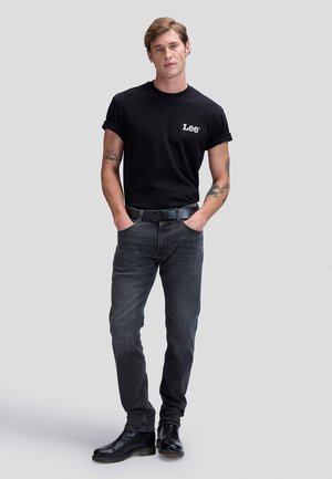 Uomo in piedi con le mani nelle tasche, indossa una t-shirt nera Lee, jeans scuri, cintura nera, stivali neri e mostra tatuaggi su entrambe le braccia.