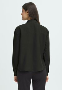 Woman with long dark hair wears a black long-sleeve shirt and black pants, standing facing away against a plain light background.