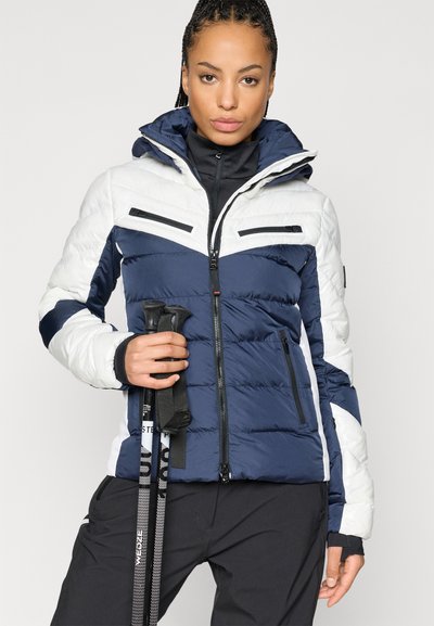Φουσκωτό μπουφάν σε ναυτικό μπλε και λευκό με σχέδιο ζιγκ-ζαγκ, που διαθέτει κουκούλα και πολλαπλές τσέπες με φερμουάρ, κρατώντας πεζοπορικά μπαάστα.