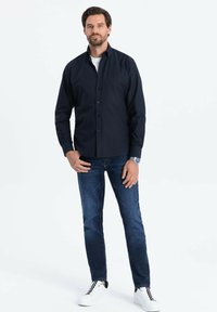Camisa azul marino de botones, ajustada, con un bolsillo. Combinada con jeans de mezclilla oscuros y zapatillas blancas con cordones negros. Diseño sencillo y moderno.