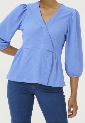 Kvinde iført en lyseblå tekstureret omslagstop med trekvart puffede ærmer og mørkeblå jeans, poserer med den ene hånd på hoften.
