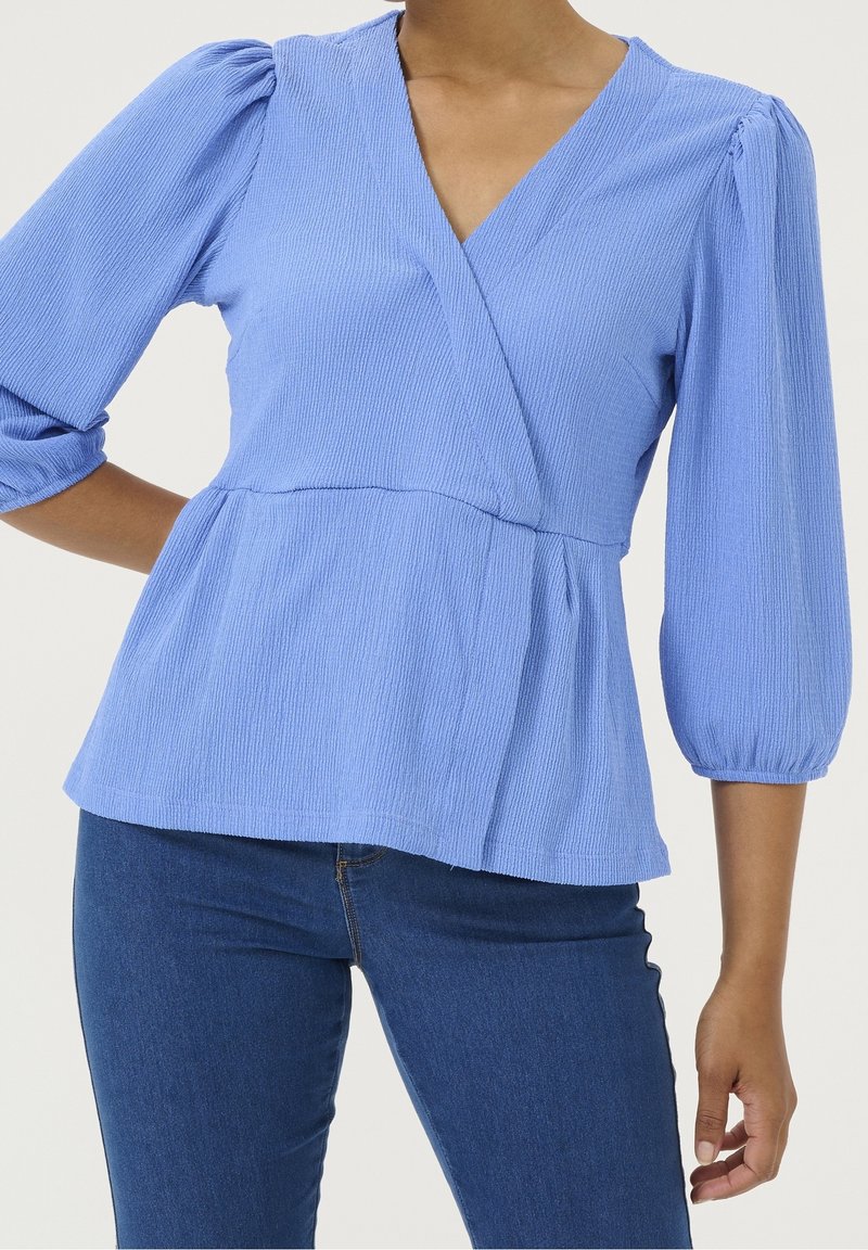 Femme portant un top cache-cœur bleu clair texturé avec des manches bouffantes trois-quarts et un jean bleu foncé, posant avec une main sur la hanche.