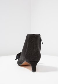 Bottine noire avec un tissu texturé, petit talon, fermeture éclair à l'arrière et un accent en nœud sur un côté.