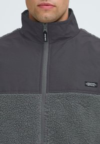 Graue Zip-Jacke mit einem Fleece-Unterteil. Verfügt über einen hohen Kragen, glattes Obermaterial und ein Markenetikett an der Tasche. Besonders ausgeprägte Texturdetails.