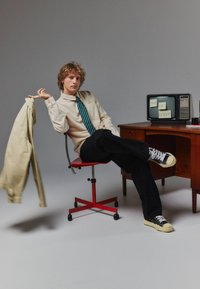 Beżowa koszula z guzikami, paskowana tealowa krawat, czarne spodnie i sneakersy. Siedzi na czerwonym krześle obok drewnianego biurka z starą telewizją wyświetlającą napis "POZA BIUREM."