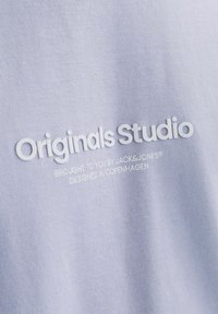 Gaiši purpura audums ar uzrakstu "Originals Studio" paaugstinātajās baltajās burtos, ar papildu tekstu mazākā fontā zem tā. Mīksta tekstūra.